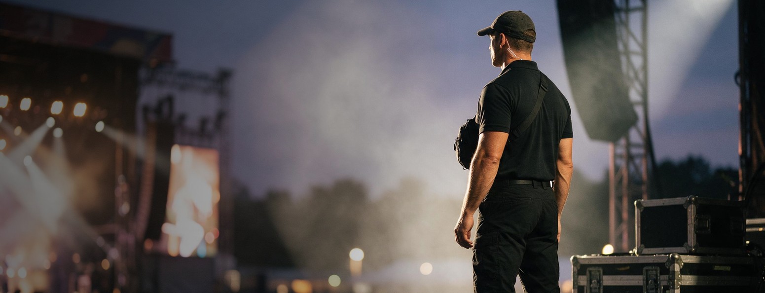 Tour and artist security: a DUSTWUN protection agent in a black cap and polo with a chest sling bag stands side-stage at a festival at dusk, watching the crowd through warm stage haze